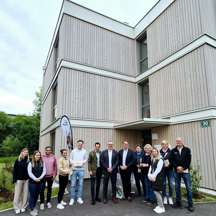 Eine Gruppe von Menschen, wahrscheinlich ein Team, steht vor einem modernen Gebäude. Einige halten Geschenke in den Händen, was auf einen festlichen Anlass hindeutet.