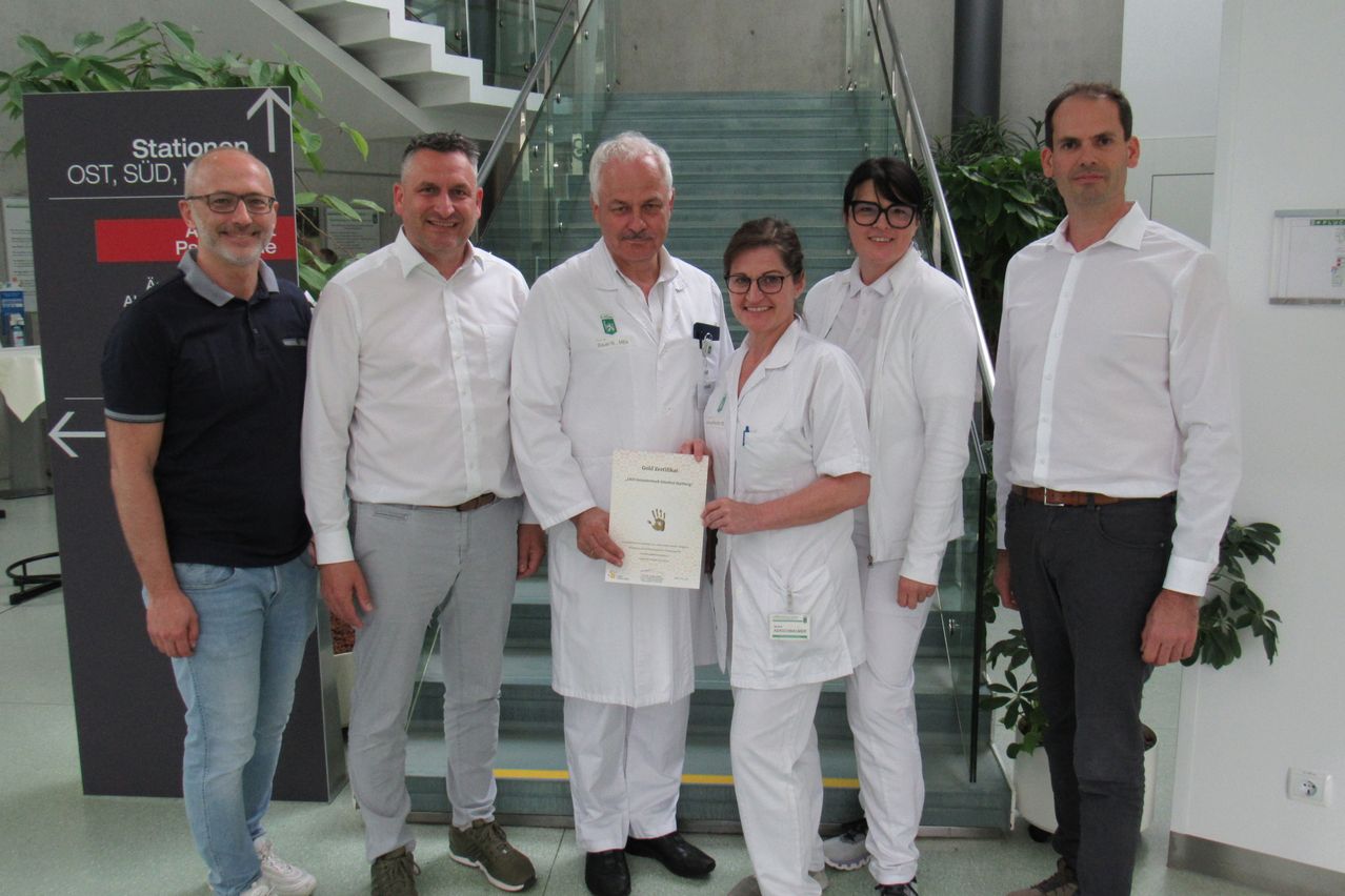 Eine Gruppe von Menschen in weißen Laborkittel steht auf einer Treppe und posiert für ein Foto. Eine Frau hält ein Zertifikat, und der Mann neben ihr hat einen Namensschild. Hinter ihnen befindet sich eine Treppe mit Glasgeländer und Pflanzen.