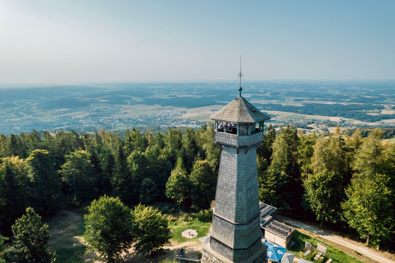 Ein hoher Turm steht auf einem Hügel, umgeben von üppigem Baumbestand. Besucher befinden sich auf dem Balkon und genießen die Aussicht. Die Landschaft erstreckt sich mit Wäldern und weit entfernten Städten unter einem klaren Himmel.