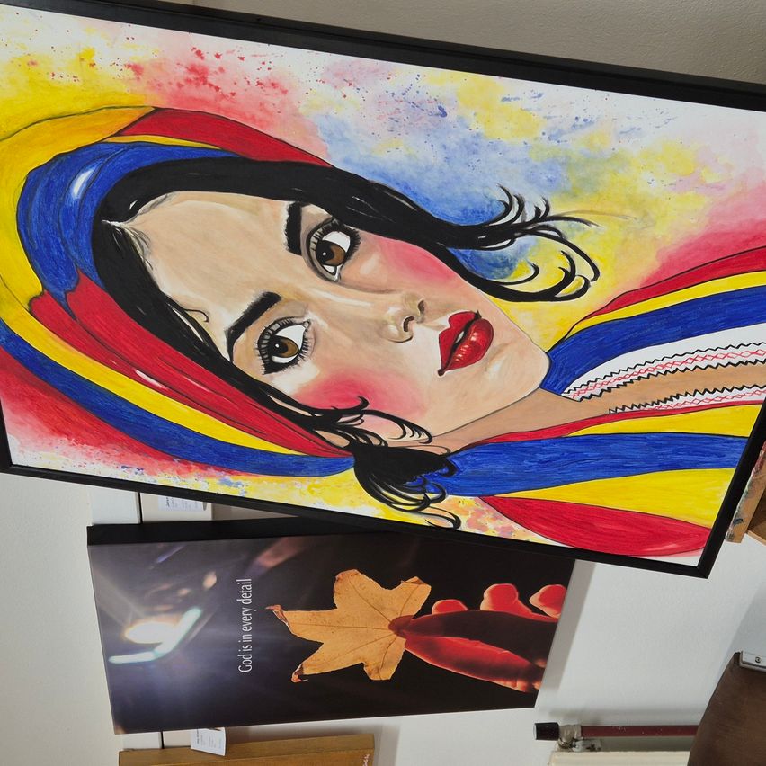 Ein farbenfrohes Gemälde einer Frau mit langen schwarzen Haaren, die einen bunten Schal trägt, hängt an einer Wand. Darunter befindet sich ein gerahmter Druck mit der Aufschrift 'Gott ist in jedem Detail.'