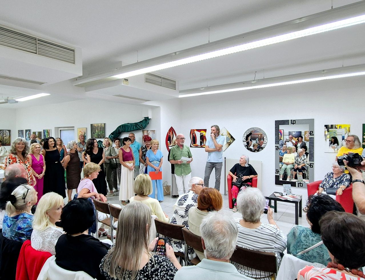 Eine Gruppe von Menschen in einem Raum, einige stehend und andere sitzend, scheint eine Kunstgalerie zu sein. Eine ältere Frau sitzt auf einem Stuhl und hält eine Handtasche. Eine Person steht vor ihnen und spricht möglicherweise. Der Raum hat weiße Wände, Deckenleuchten und einen Spiegel.