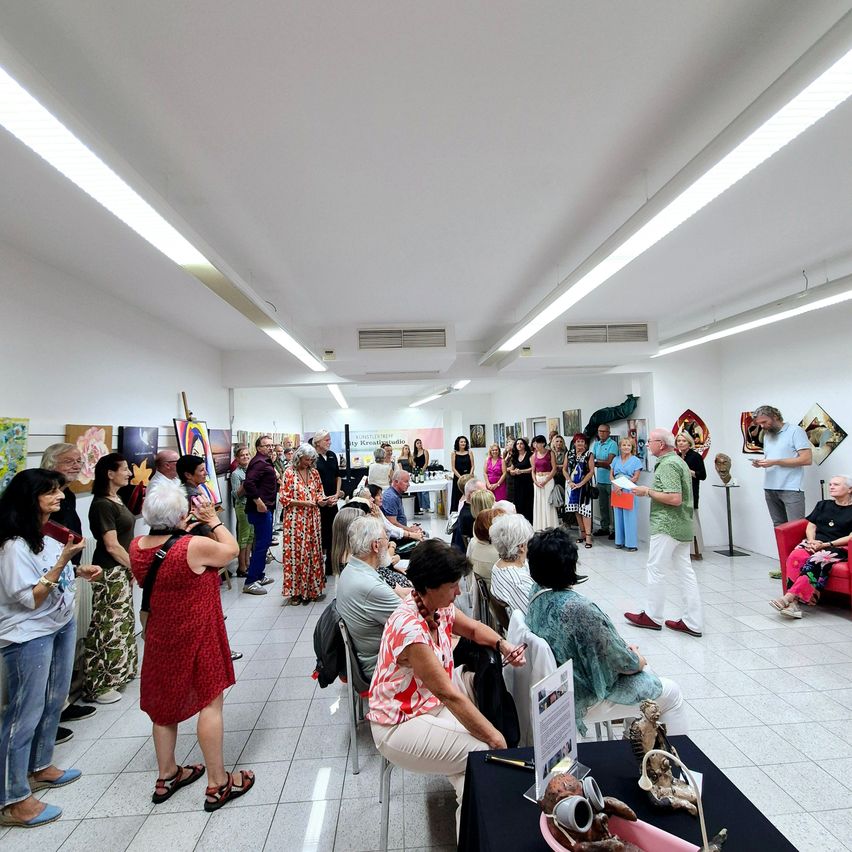 Eine Gruppe von Menschen befindet sich in einer Kunstgalerie. Einige stehen und andere sitzen. Es gibt Gemälde an den Wänden. Ein Mann in einem grünen Hemd spricht die Menge an.