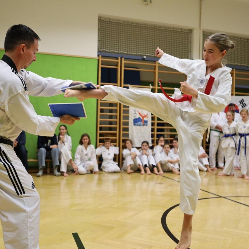 Ein Mann und ein Mädchen in weißen Uniformen üben Karate. Der Mann hält ein Buch, und das Mädchen tritt. Andere Kinder beobachten.