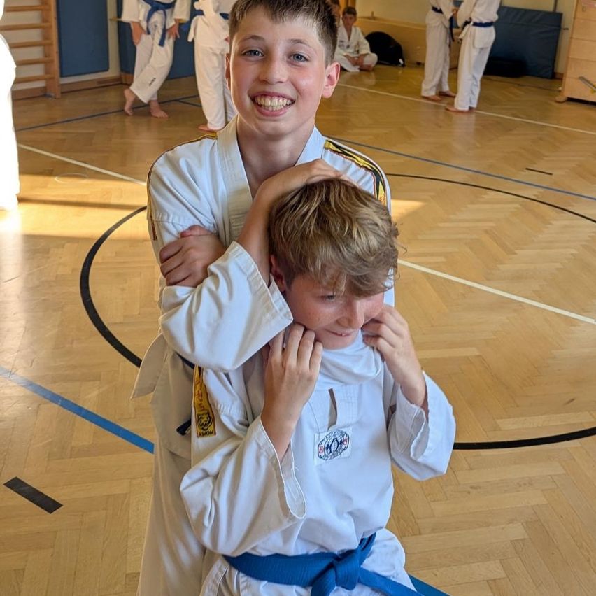Zwei junge Jungen in Kampfsportuniformen führen ein spielerisches Ringen in einer Turnhalle aus. Der Junge im Vordergrund scheint zu kämpfen, während der andere Junge lächelt. Im Hintergrund üben andere auf Matten.