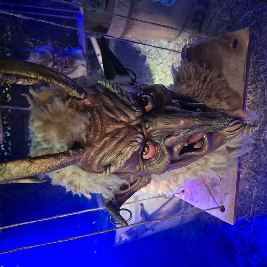 Ein teuflisch aussehender Kopf mit Hörnern, großen Augen und einem aufgerissenen Mund hängt an Fäden. Der Kopf hat weißes Fell um Gesicht und Hals.