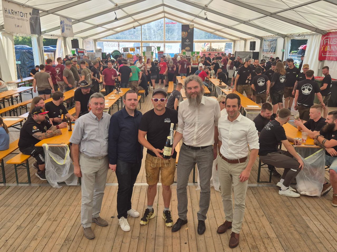 Fünf Männer stehen in einem überfüllten Bierzelt und lächeln. Einer hält eine Flasche Bier. Sie sind von langen Tischen und vielen Menschen umgeben, die ein Festival genießen.