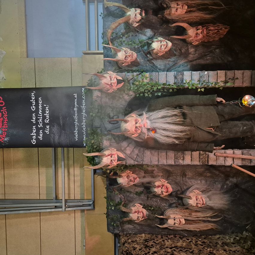 Ein Werbedisplay für 'Mutter der Gruten' zeigt eine Reihe unheimlicher, gehörnter Gesichter vor einem dunklen Hintergrund. Im Vordergrund liegt eine Figur, umrankt von Pflanzen und Ranken.