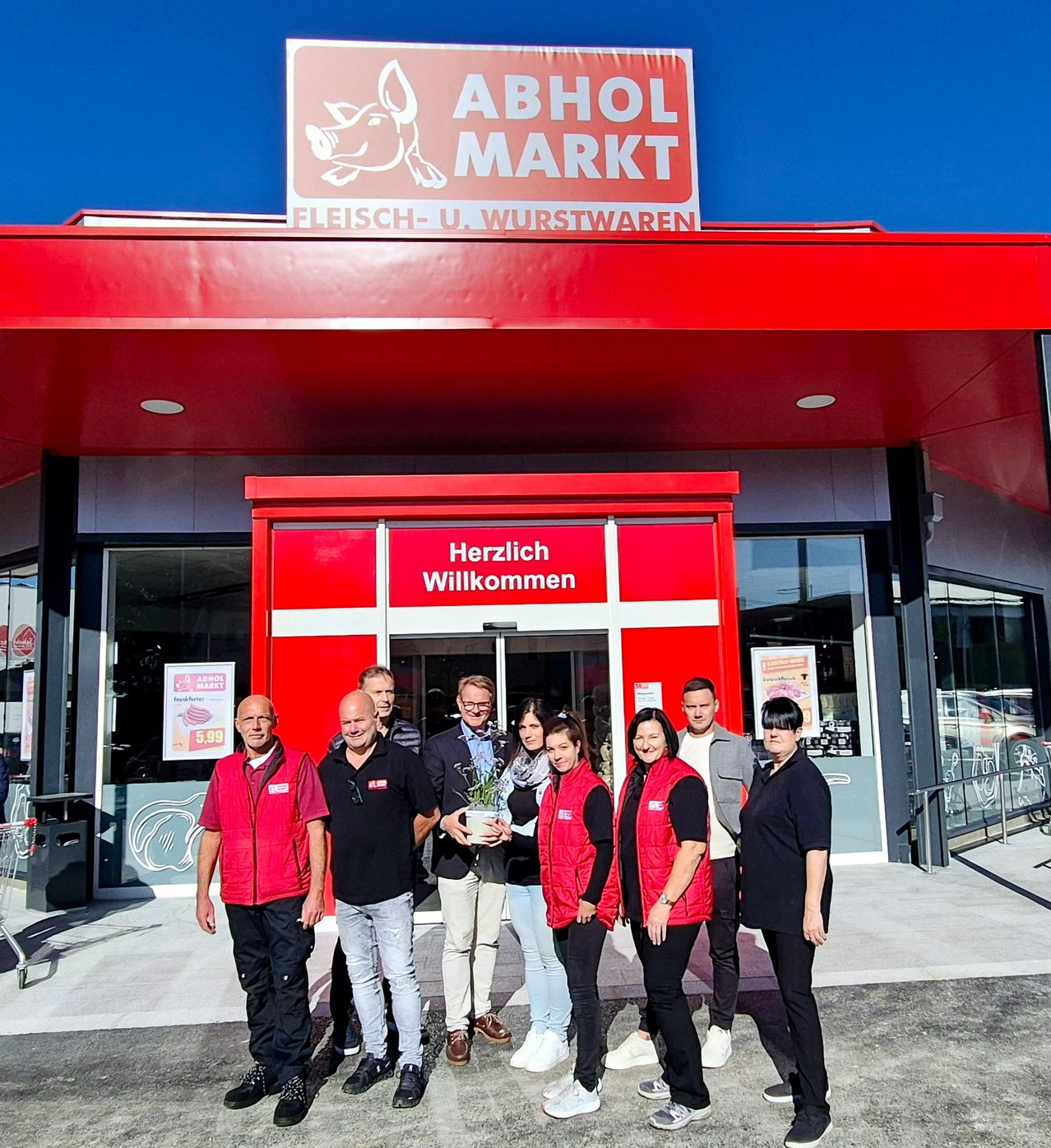 Eine Gruppe von Menschen steht lächelnd vor einem Geschäft. Das Geschäft hat ein rotes Dach und das Schild sagt 'ABHOL MARKT Fleisch- und Wurstwaren.' Die Tür hat die Aufschrift 'Herzlich Willkommen' und sie halten eine Pflanze.