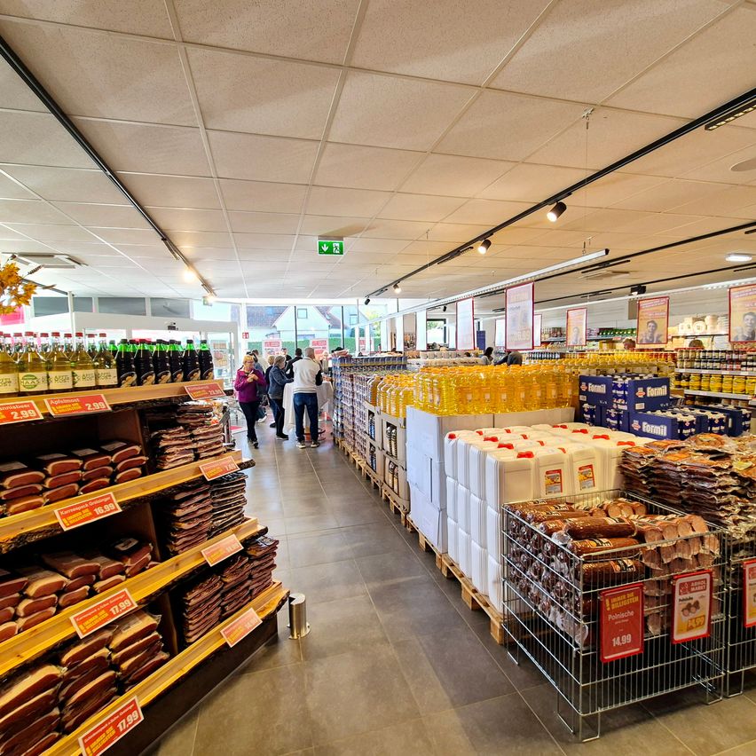Ein Supermarktregal zeigt eine Vielzahl von Produkten, darunter Fleisch, Öle und verpackte Waren, mit Kunden beim Stöbern. Die Decke hat Beleuchtung und Schilder.
