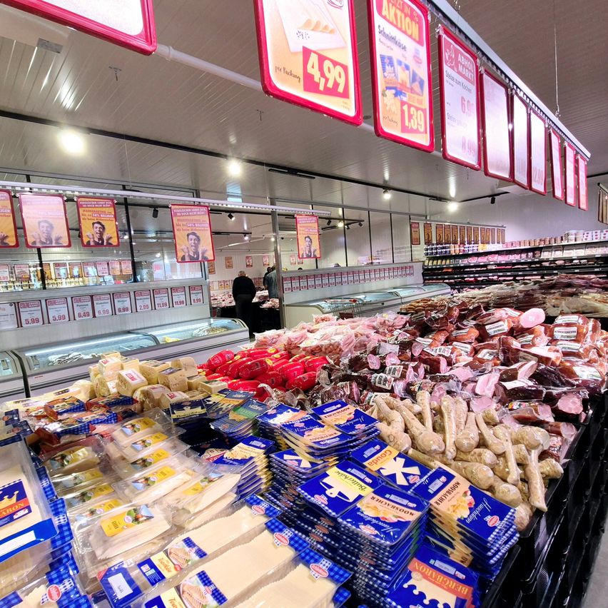 Ein Lebensmittelgeschäft mit verschiedenen Lebensmitteln zur Schau, einschließlich Käse, Würsten und Fleisch, alles ordentlich angeordnet mit sichtbaren Preisschildern. Es gibt auch Werbeplakate, die von der Decke hängen.