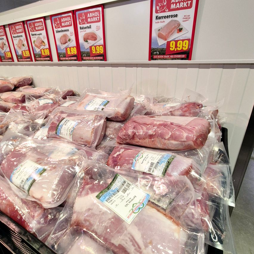 Eine Fleischausstellung mit mehreren Fleischpaketen in Plastik. Das Fleisch ist mit Preisschildern und Namen beschriftet. Der Hintergrund zeigt eine weiße Wand mit roten Postern, die Fleisch bewerben.