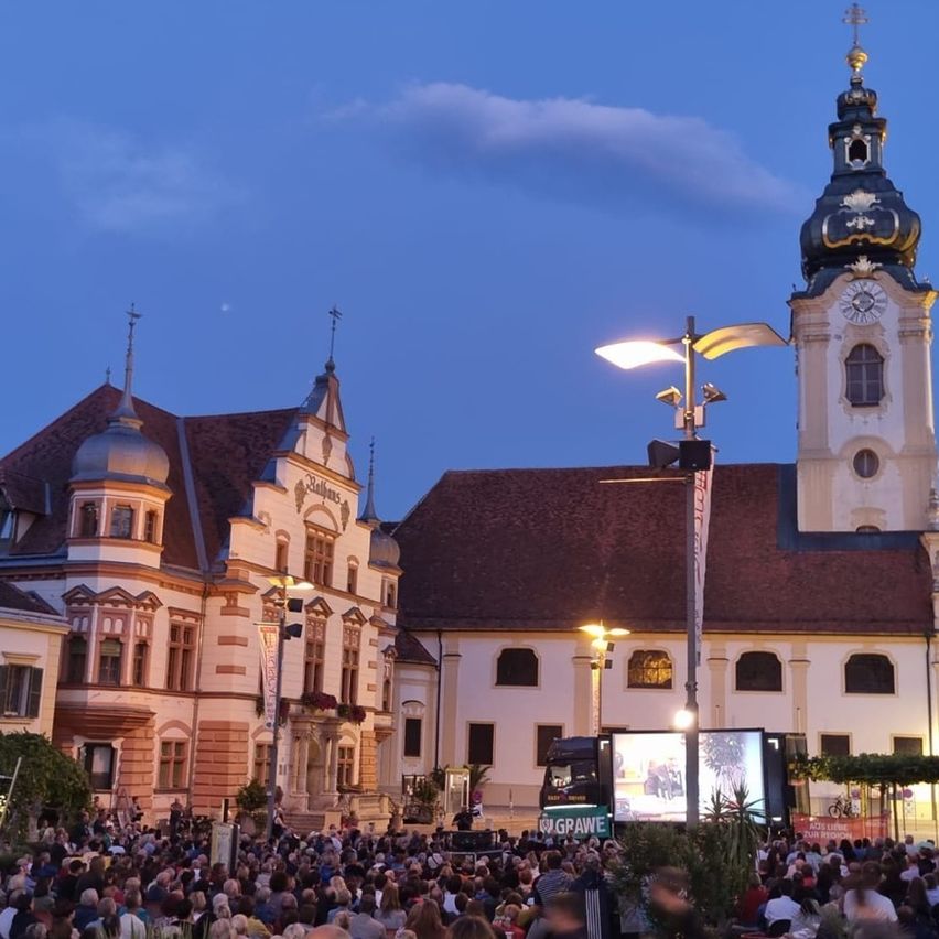 Eine Menschenmenge versammelt sich vor einem architektonischen Gebäude mit Turm und Uhr. Der Himmel ist klar mit Mond und Wolken. Es gibt Straßenlaternen und eine Leinwand auf einem Lastwagen.