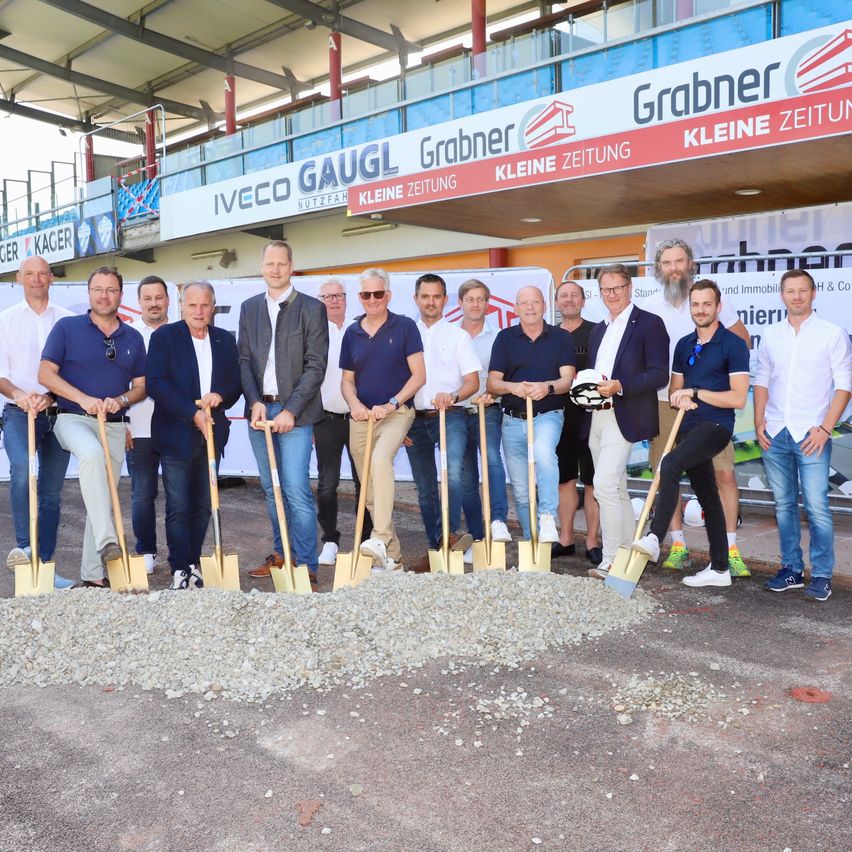 Eine Gruppe von Männern steht auf einer Baustelle und hält Schaufeln in der Hand, wahrscheinlich bei einer Grundsteinlegung. Im Hintergrund sind Banner mit den Texten 'Grabner Kleine Zeitung' und 'IVECO GAUGL' zu sehen.