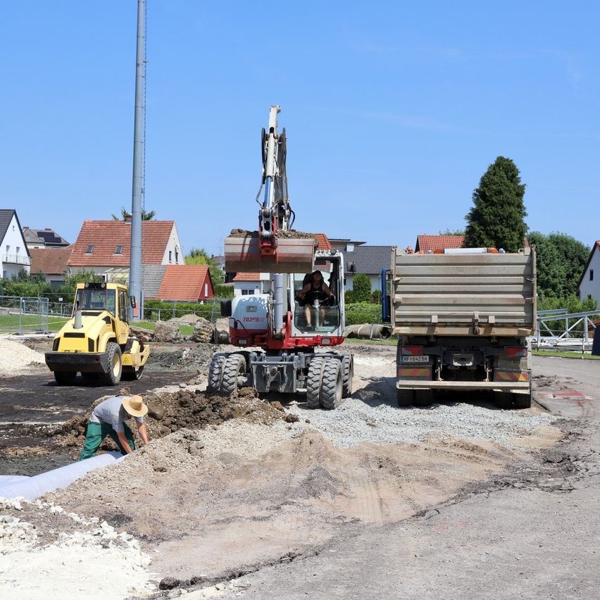 Auf einer Baustelle werden zwei schwere Fahrzeuge, ein Kran und ein Lastwagen, eingesetzt. Ein Mann legt eine Rohrleitung auf dem Boden.
