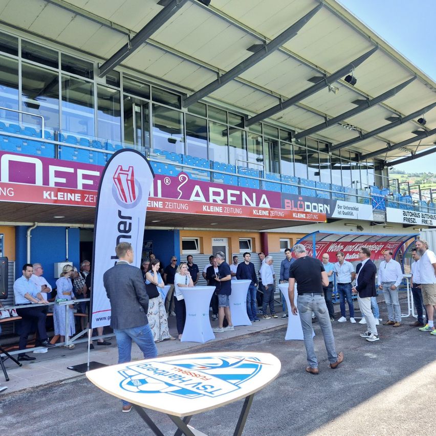 Eine Gruppe von Menschen steht vor einer Arena mit Bannern und einem Tisch im Vordergrund. Die Arena hat Glasfenster und blaue Sitze.
