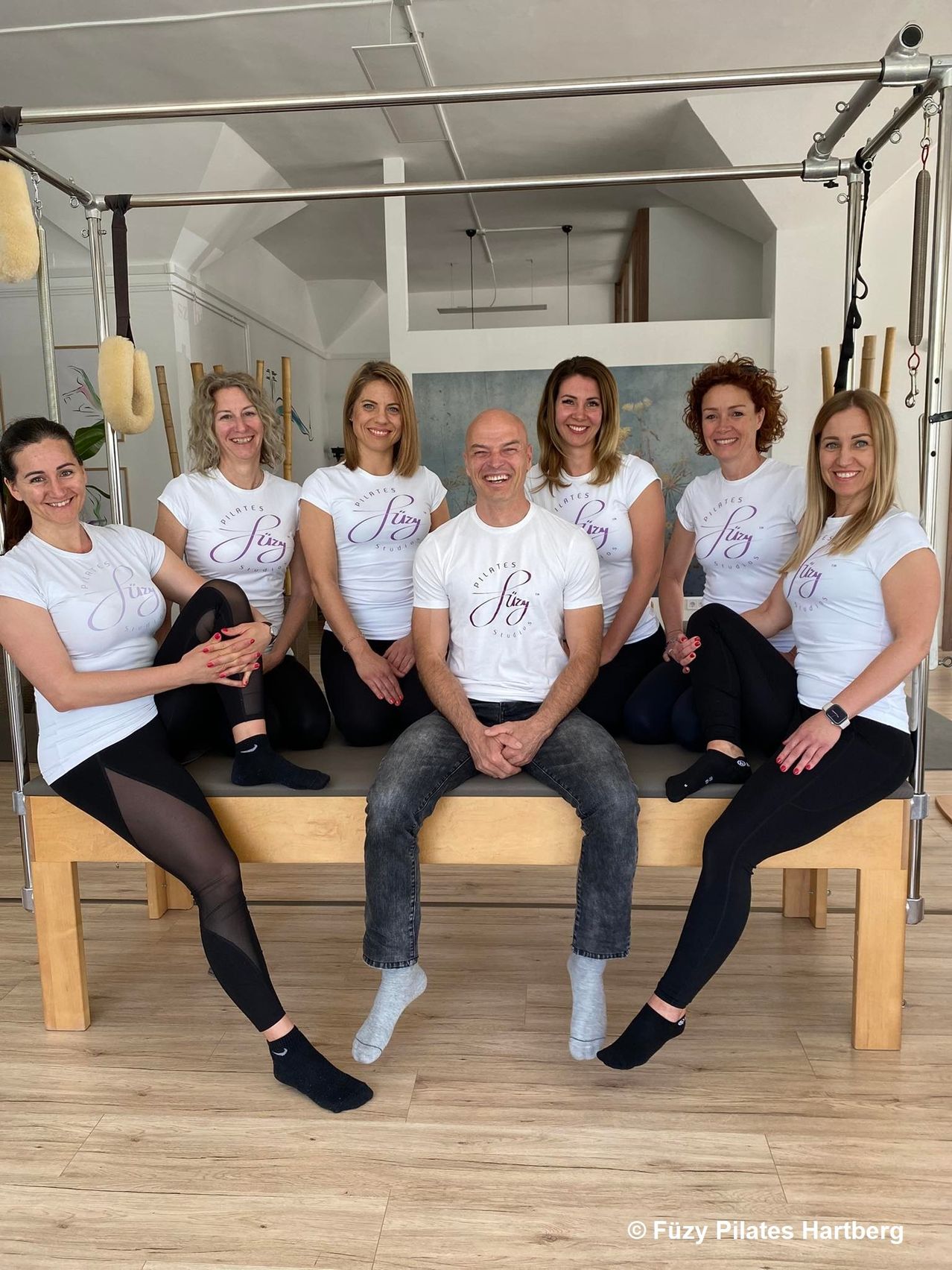 Eine Gruppe lächelnder Frauen und ein Mann in Trainingskleidung sitzen auf einer Bank, alle tragen weiße T-Shirts mit den Worten 'Pilates' und 'Sty' in Lila. Sie scheinen sich in einem Studio-Setting zu befinden, mit Trainingsgeräten im Hintergrund sichtbar.