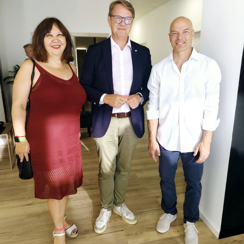 Drei Menschen stehen lächelnd auf einem Holzboden für ein Foto. Eine Frau in einem roten Kleid, ein Mann in einem blauen Blazer und ein anderer Mann in einem weißen Hemd. Dahinter eine weiße Wand und eine Topfpflanze.