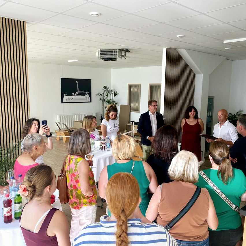 Eine Gruppe von Menschen, darunter zwei Männer und mehrere Frauen, steht in einem Raum mit einem Tisch in der Mitte. Eine Frau macht ein Foto mit ihrem Handy. Es gibt ein Bild an der Wand und eine Pflanze in der Ecke.
