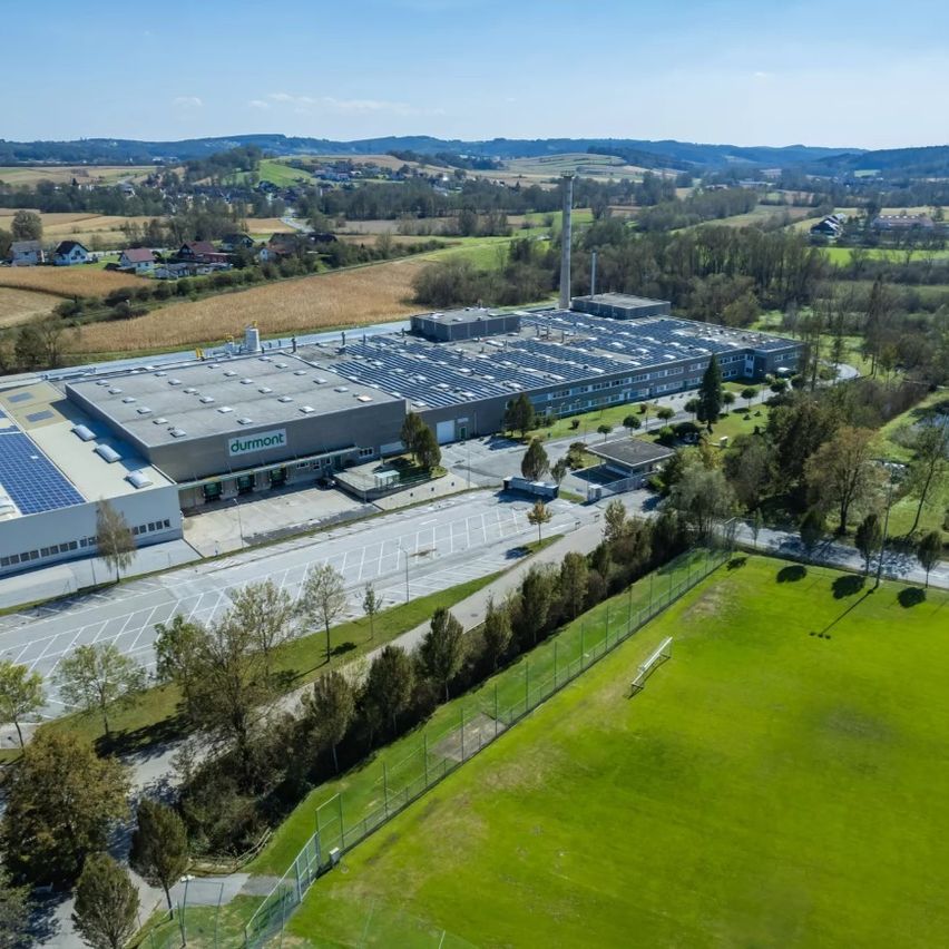 Ein Luftbild einer großen Fabrik mit Solarpanelen auf dem Dach. Die Fabrik ist von einem weitläufigen, offenen Feld mit einem Zaun und einem Rasen umgeben. Im Vordergrund sind Bäume und Büsche zu sehen.