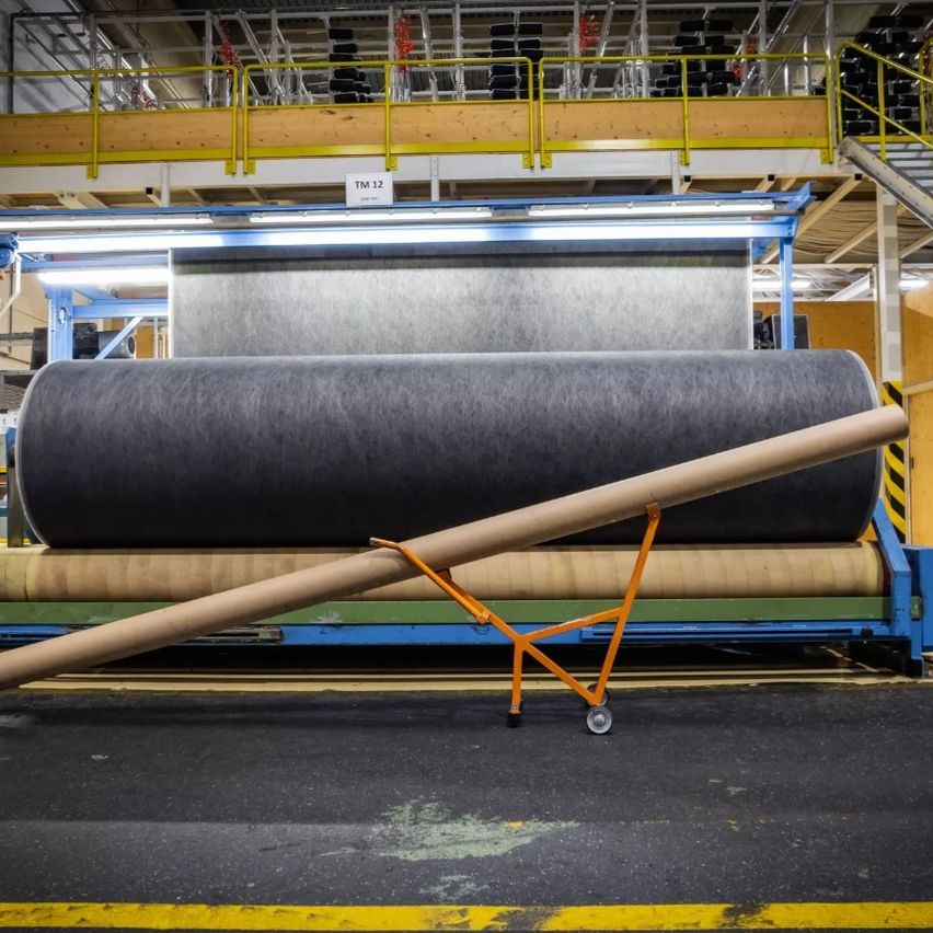 Eine große Rolle Stoff wird in einer Fabrik auf einem Förderband bewegt. Sie wird von einem orangefarbenen Wagen mit Rädern unterstützt. Ein weißes Schild mit 'TM 12' ist darüber montiert.