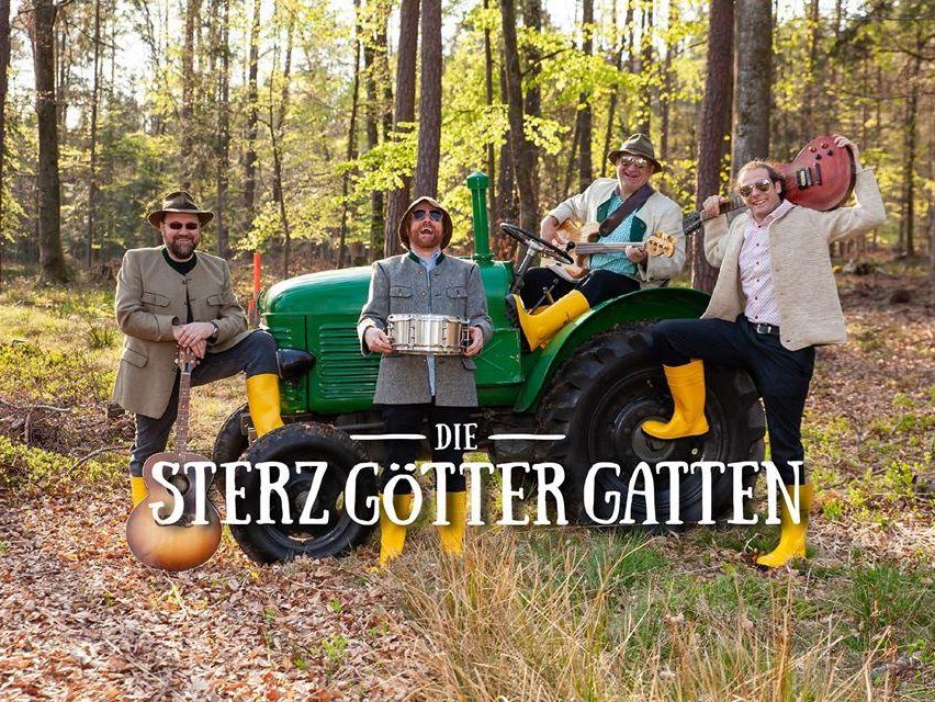 Eine Band von vier Männern posiert in einem Wald mit einem Traktor, ein Mann sitzt auf dem Traktor mit einer Gitarre, ein anderer hält eine Trommel und zwei andere stehen mit Gitarren.