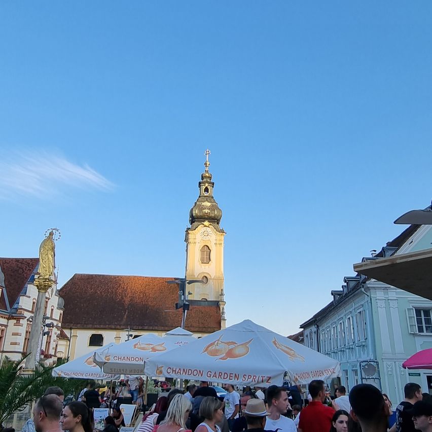 Eine Menschenmenge versammelt sich um einen Markt mit weißen Sonnenschirmen, einem prominenten Kirchturm im Hintergrund und einem blauen Himmel.