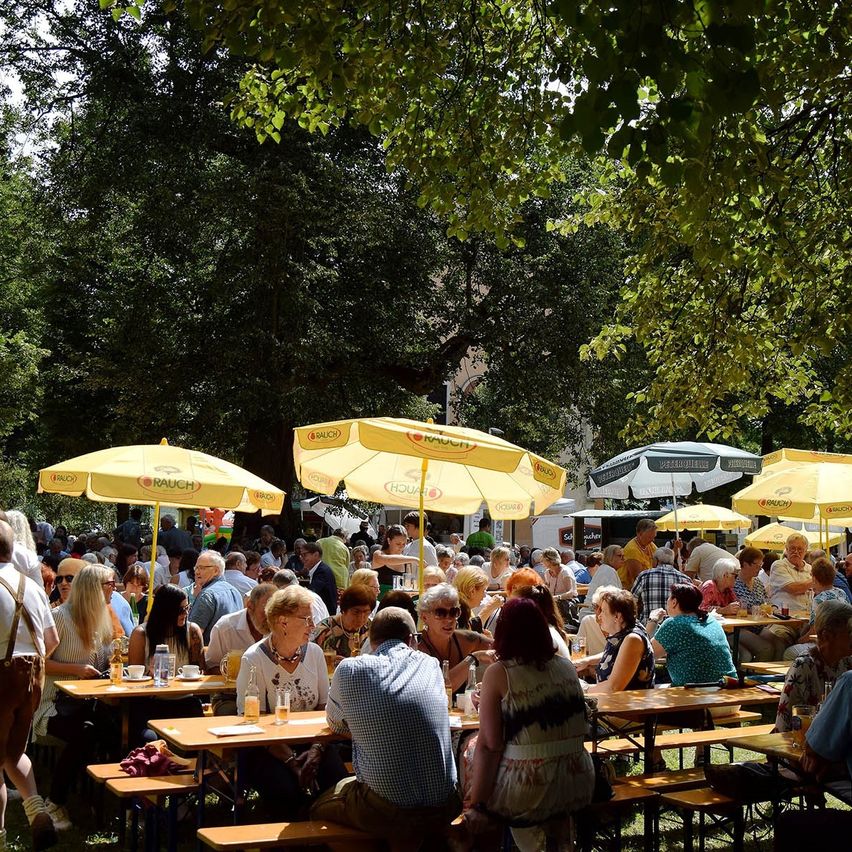 Ein Freiluft-Essbereich mit vielen Menschen, die an Tischen unter Schirmen sitzen. Die Tische sind mit Essen und Getränken beladen. Bäume spenden im Hintergrund Schatten.