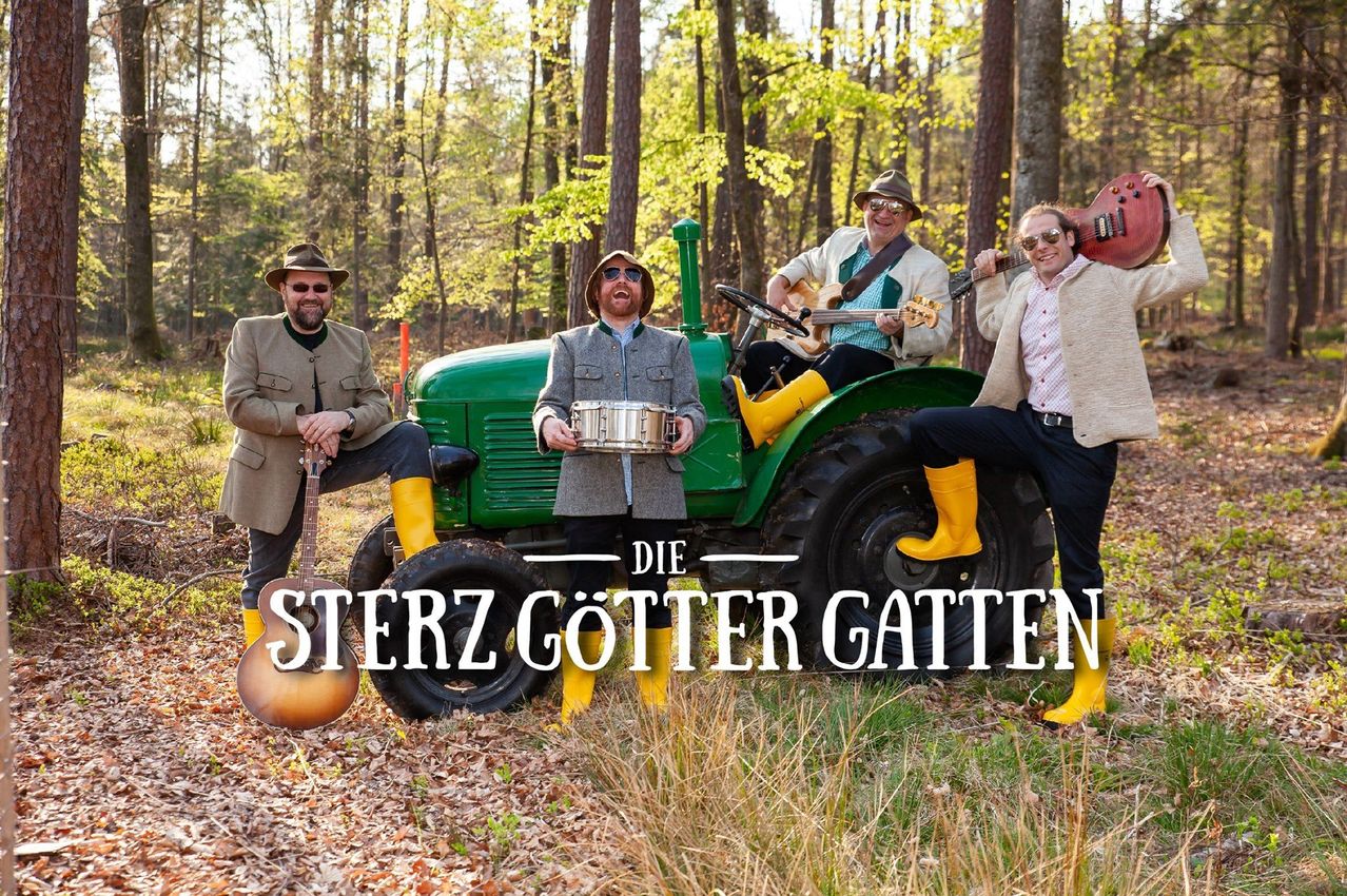 Vier Männer stehen um einen grünen Traktor im Wald herum, jeder hält ein Musikinstrument. Ein Mann sitzt auf dem Traktor und hält eine Gitarre, während ein anderer steht und eine Trommel hält. Die Worte 'Die Sterz Götter Gatten' sind in Weiß über das Bild geschrieben.