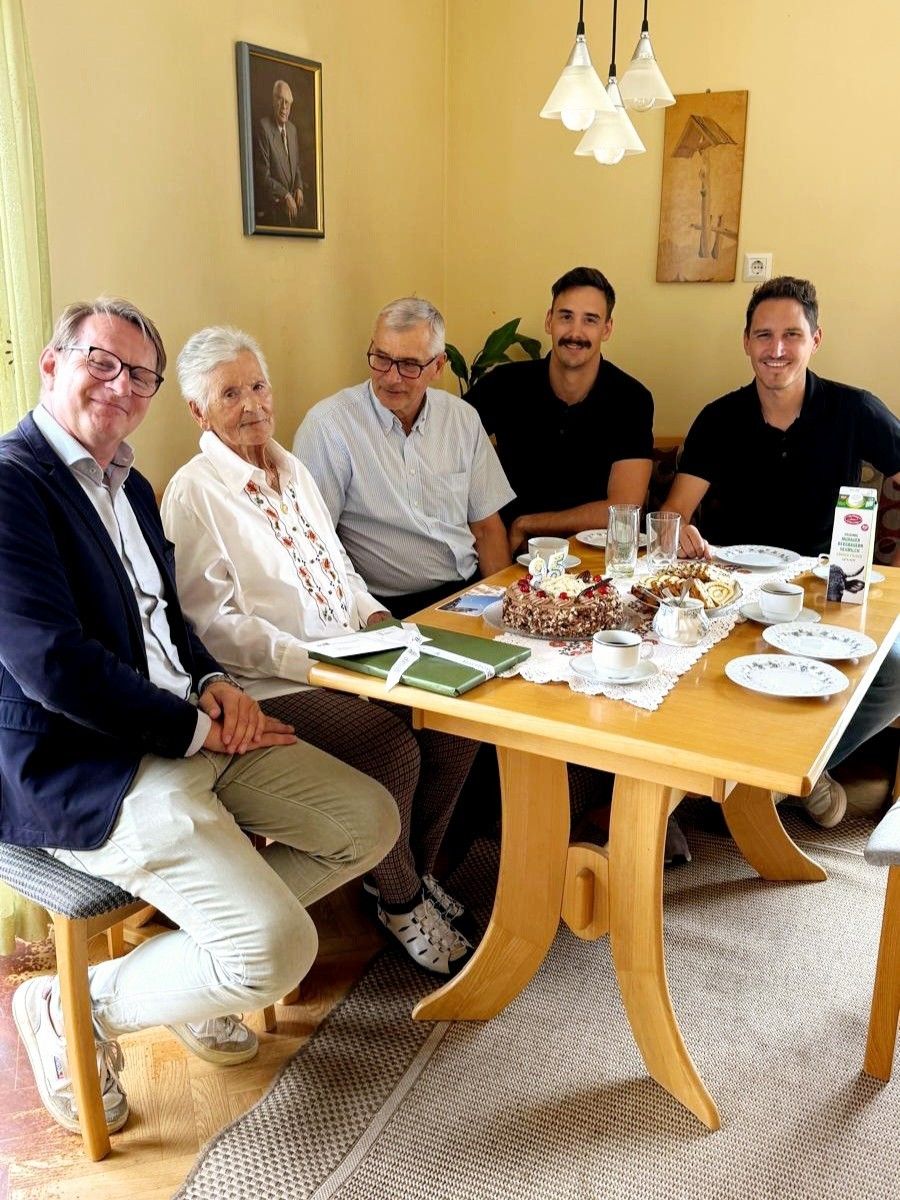 Fünf Personen sitzen lächelnd um einen Tisch herum und posieren für ein Foto. Der Tisch hat einen Kuchen, Tassen und Teller. Der Hintergrund zeigt eine gelbe Wand mit einem Bilderrahmen und einer Topfpflanze.