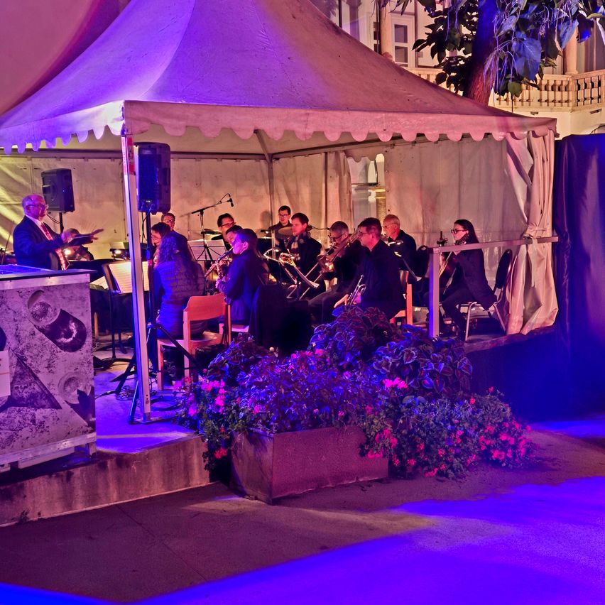 Ein Orchester spielt unter einem Zelt mit lila Beleuchtung. Die Menschen sitzen, spielen verschiedene Instrumente und ein Dirigent leitet die Aufführung.