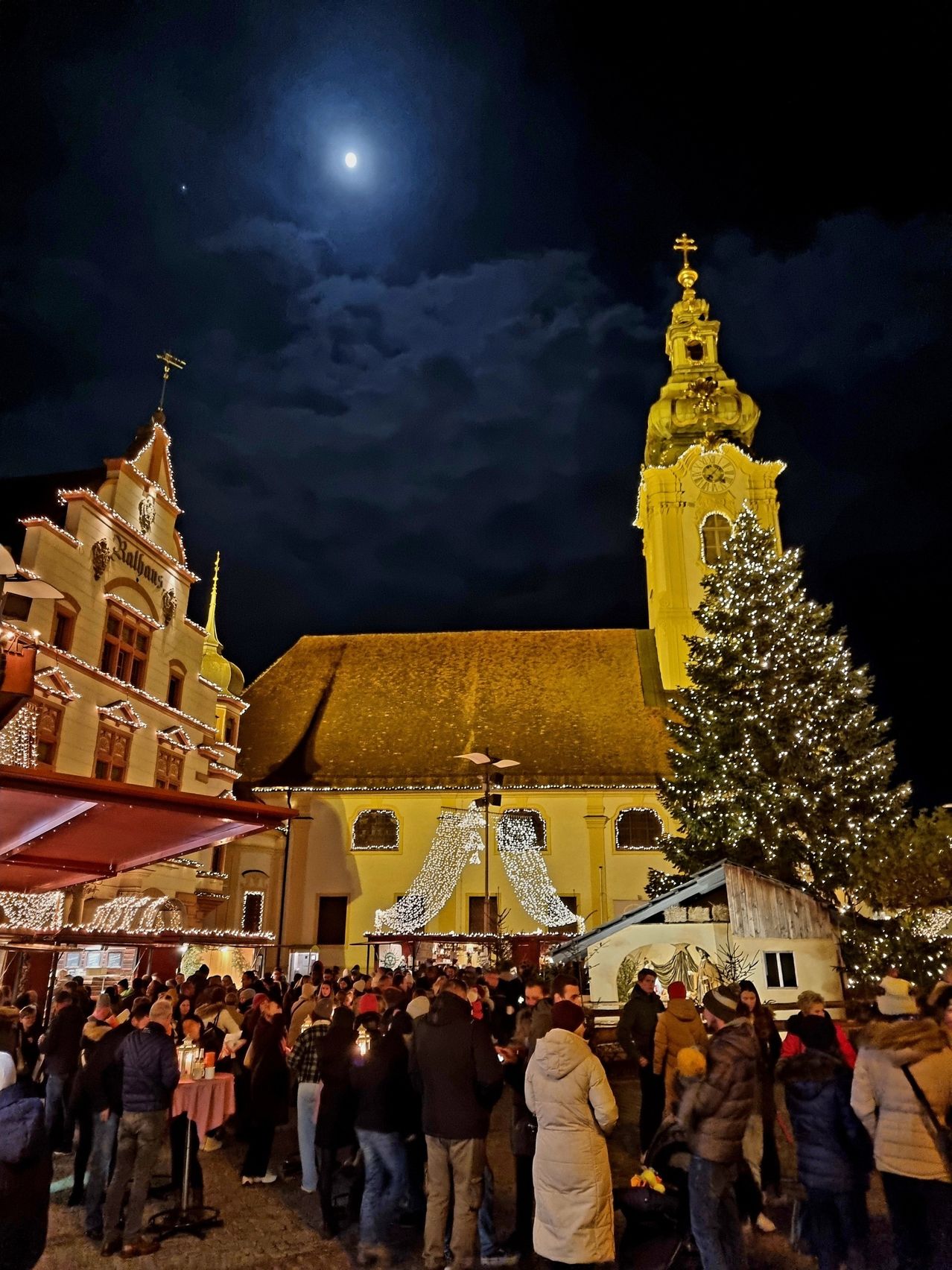 Eine Menschenmenge versammelt sich bei Nacht vor einer historischen Kirche, die mit Weihnachtslichtern und einem großen Baum geschmückt ist.