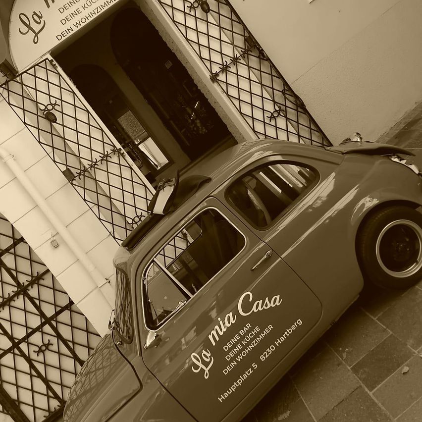 Ein Oldtimer mit der Aufschrift 'La mia Casa' parkt vor einem Gebäude mit einem Metalltor. Das Gebäude hat ein Schild mit deutschem Text.