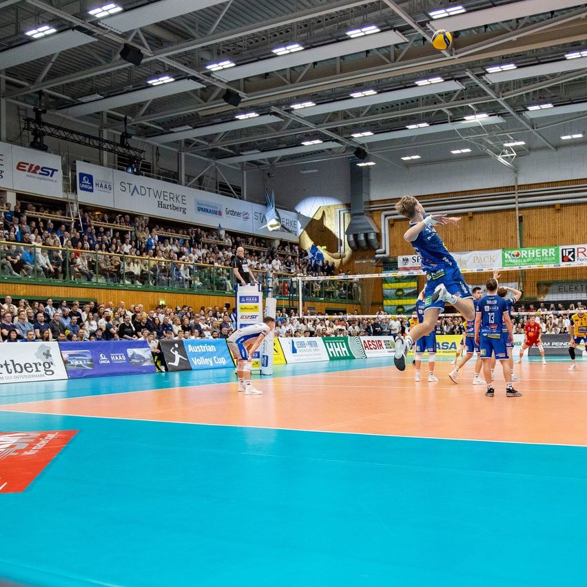 A volleyball player jumps to spike the ball. The match is taking place in an indoor stadium with many spectators.