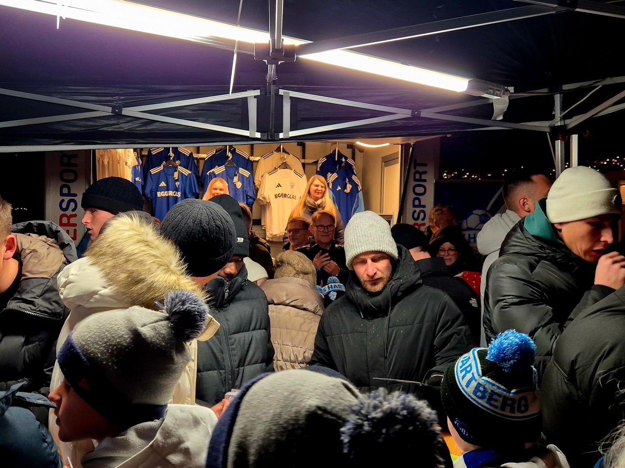 Eine Menschenmenge in Winterkleidung versammelt sich unter einem Zelt, mit ausgestellten Trikots. Ein Mann in einer schwarzen Jacke spricht die Menge an.