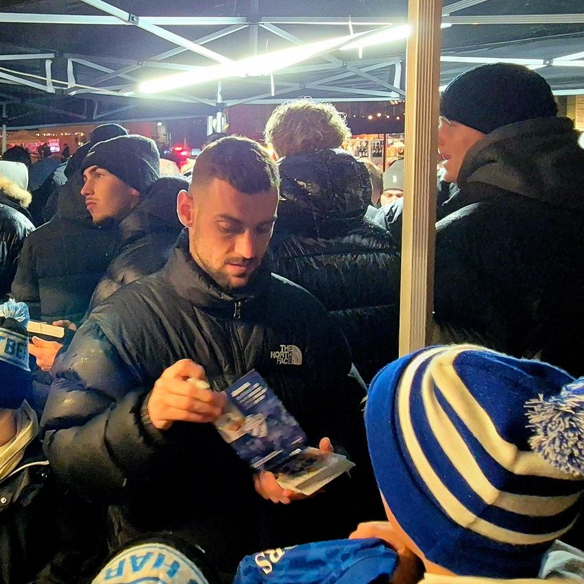 Ein Mann in einer North Face Jacke signiert Autogramme für Fans bei einer Veranstaltung, während andere in der Schlange warten.