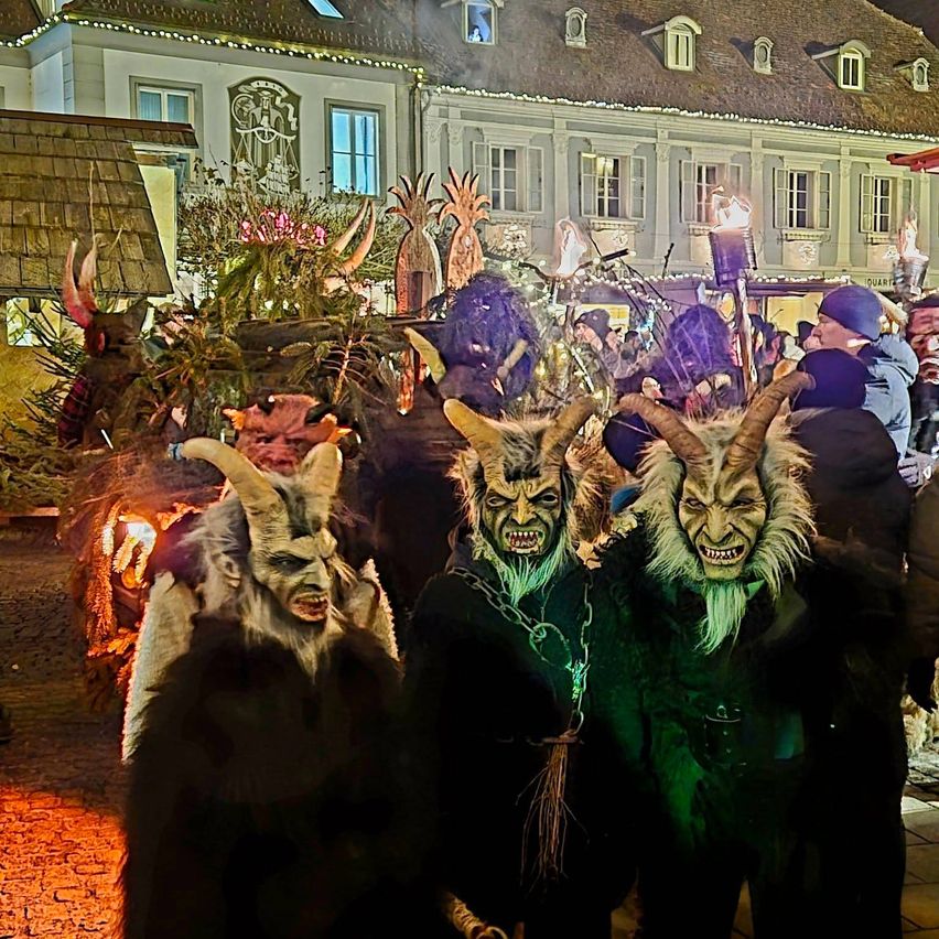 Eine Gruppe von Menschen in Hörnermasken und Pelzkostümen versammelt sich in einer festlichen Straßenszene, mit Gebäuden und festlichen Lichtern im Hintergrund.