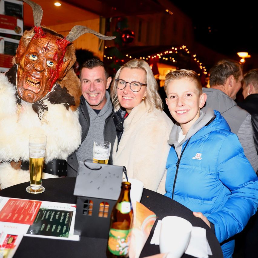 A group of people are gathered around a table at night. A man in a goat mask sits next to a smiling man and woman. They all have drinks in front of them. In the background, there are Christmas decorations.