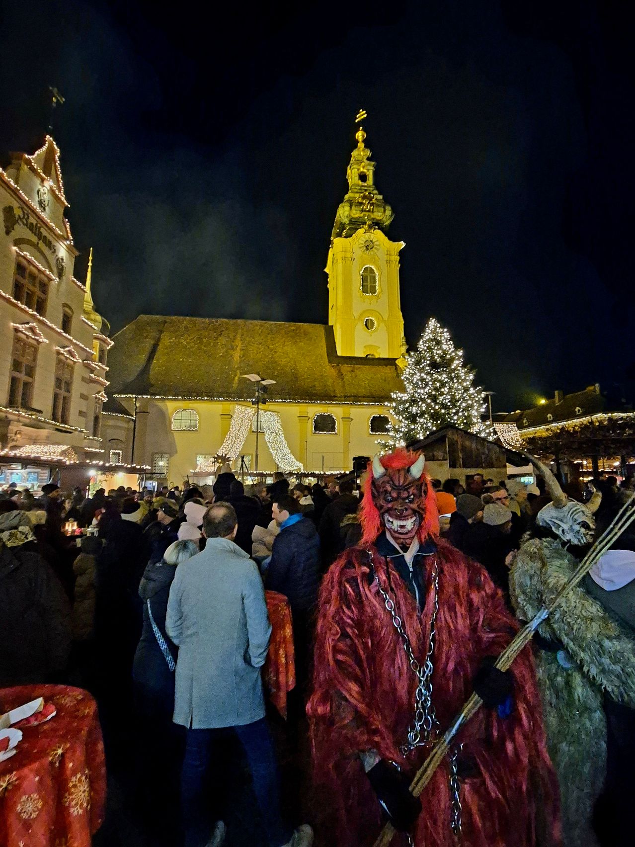 A crowd gathers in front of a church at night. The church is decorated with lights and a Christmas tree. A person in a demon costume is holding a stick and standing in the crowd.