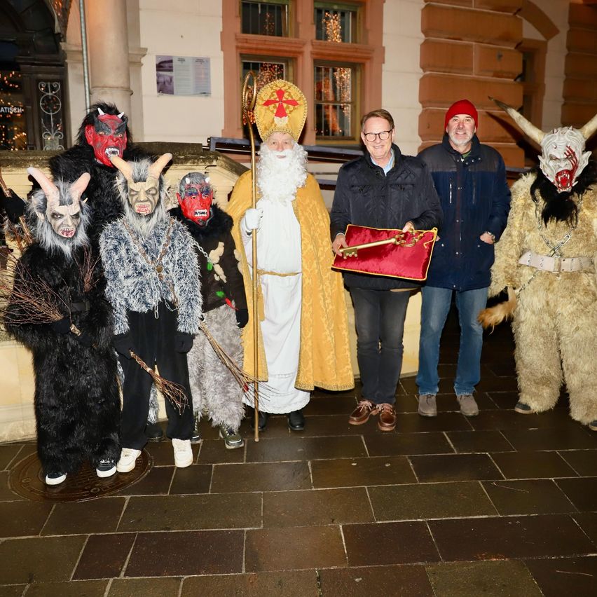 A group of people dressed in various costumes stand on a brick floor. One man holds a red bag with a key. Another man dressed as a saint holds a staff. Behind them, a building with Christmas decorations is visible.