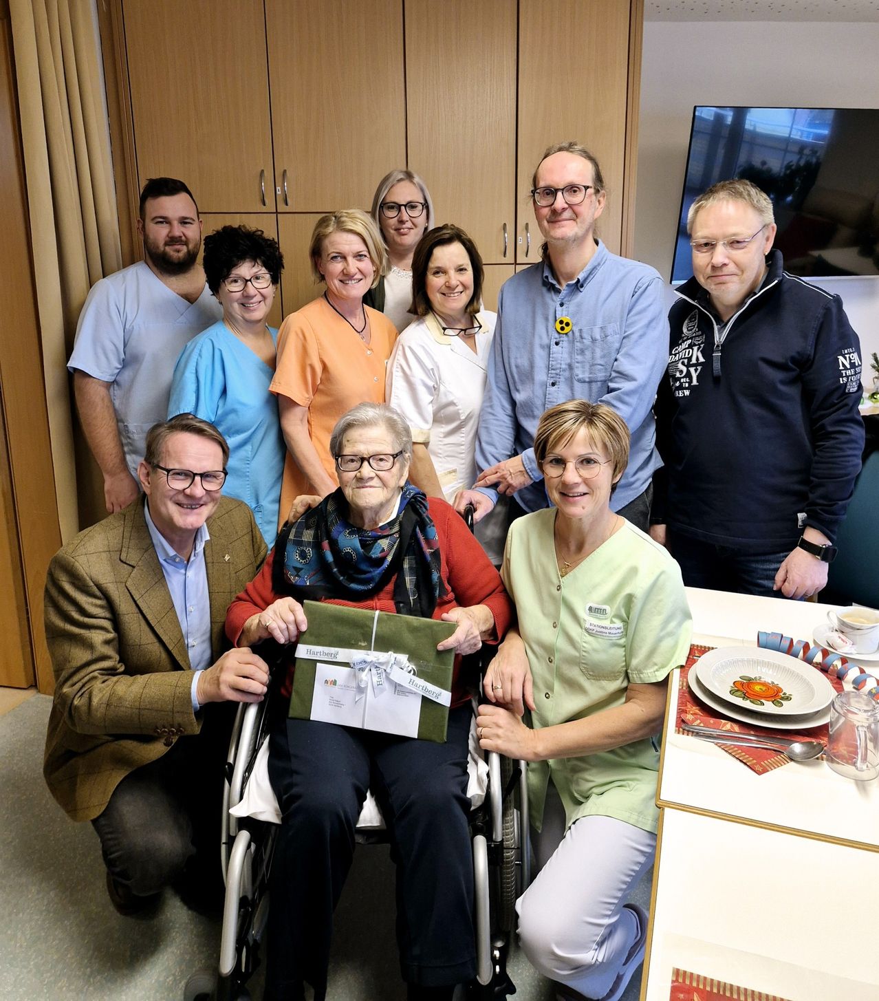 A group of people, including medical staff and visitors, are gathered in a room, posing for a photo with an elderly woman in a wheelchair receiving a gift.