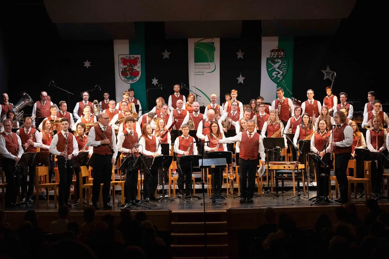 A band is performing on stage with various instruments. Some players stand, others sit. Behind them are music stands. Banners and a coat of arms are on the stage backdrop.