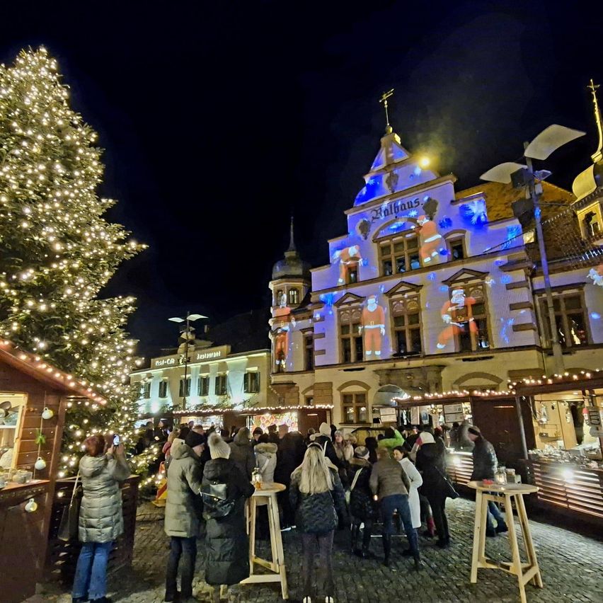 A crowd of people gathered around a decorated Christmas tree, in front of a building with festive lights and a Santa Claus projection at night.