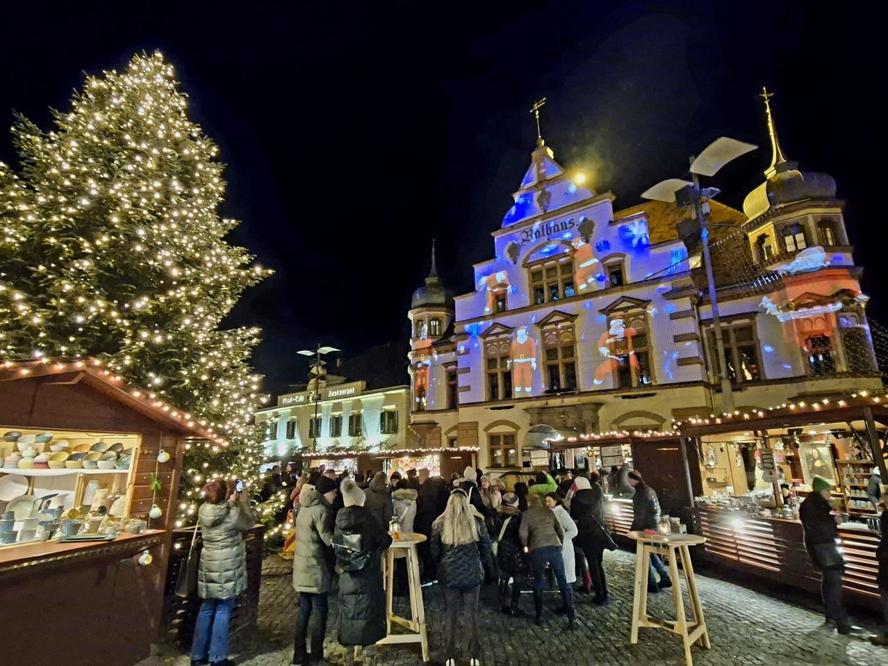 People gather around a decorated Christmas tree in front of a building with a tower, spire, and blue lights. The area is filled with festive lights and decorations.