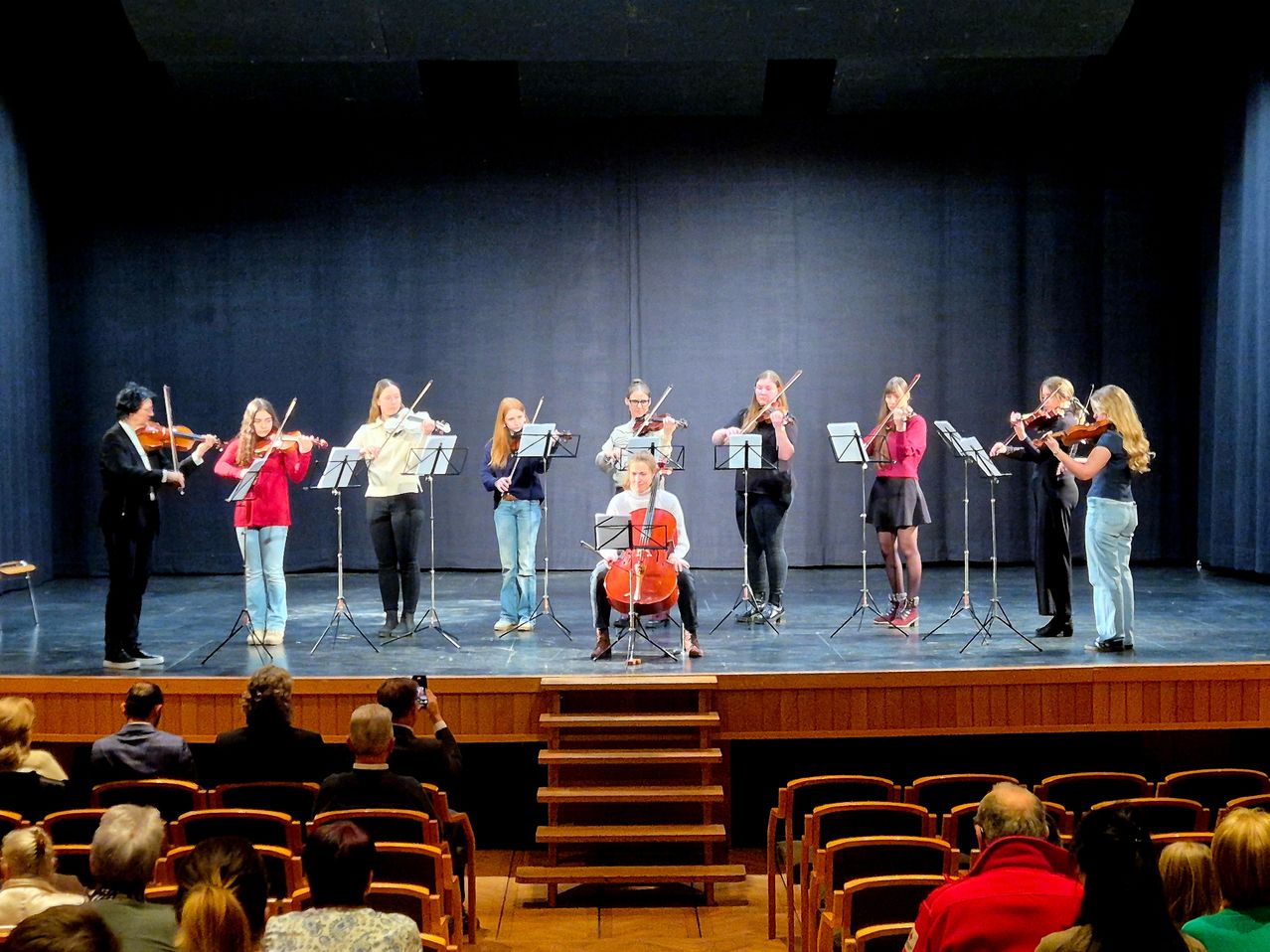A string orchestra performs on stage, with violinists and a cellist under a blue curtain. An audience watches from chairs.