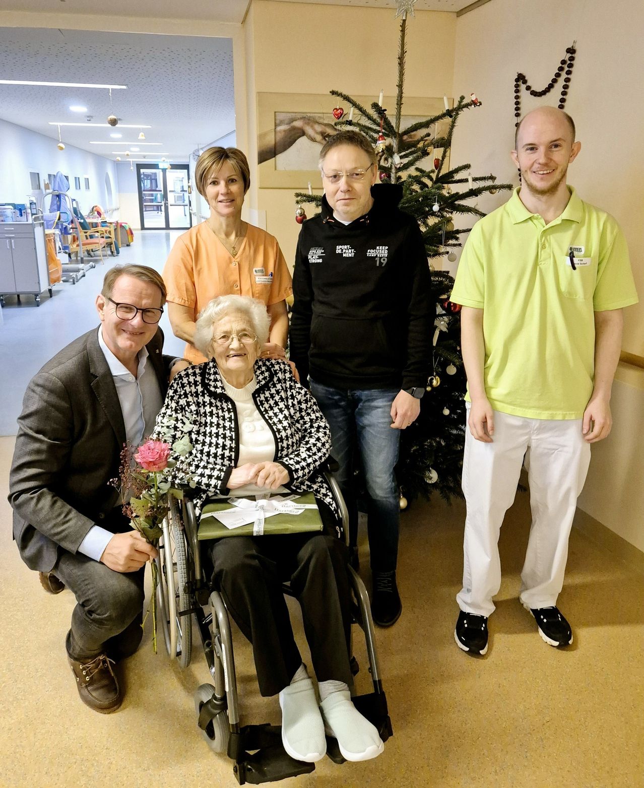 Five people pose for a picture inside a hospital. An elderly woman sits in a wheelchair, smiling. The man on her left holds a bouquet of flowers. Behind them, a Christmas tree and a woman in an orange uniform are visible.