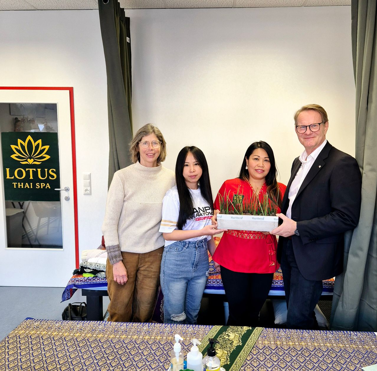 Vier Personen stehen in einem Raum mit einem Schild, auf dem Lotus Thai Spa steht. Zwei Frauen halten eine Topfpflanze. Die Frau auf der rechten Seite trägt ein rotes Oberteil.