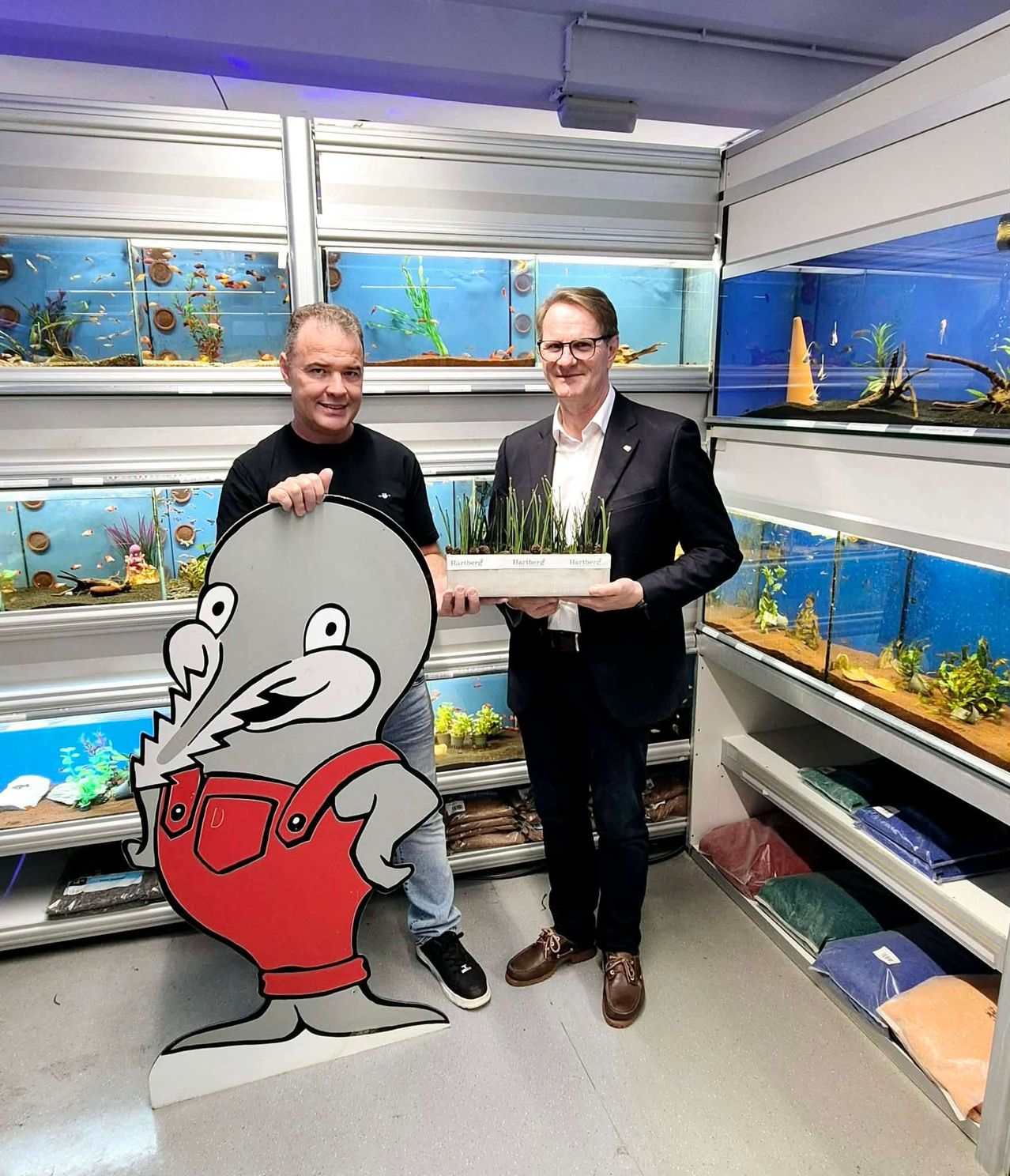 Zwei Männer stehen in einem Raum voller Aquarien, einer hält eine Pflanze und lächelt, der andere mit einer großen Nagetier-Ausschnitt.