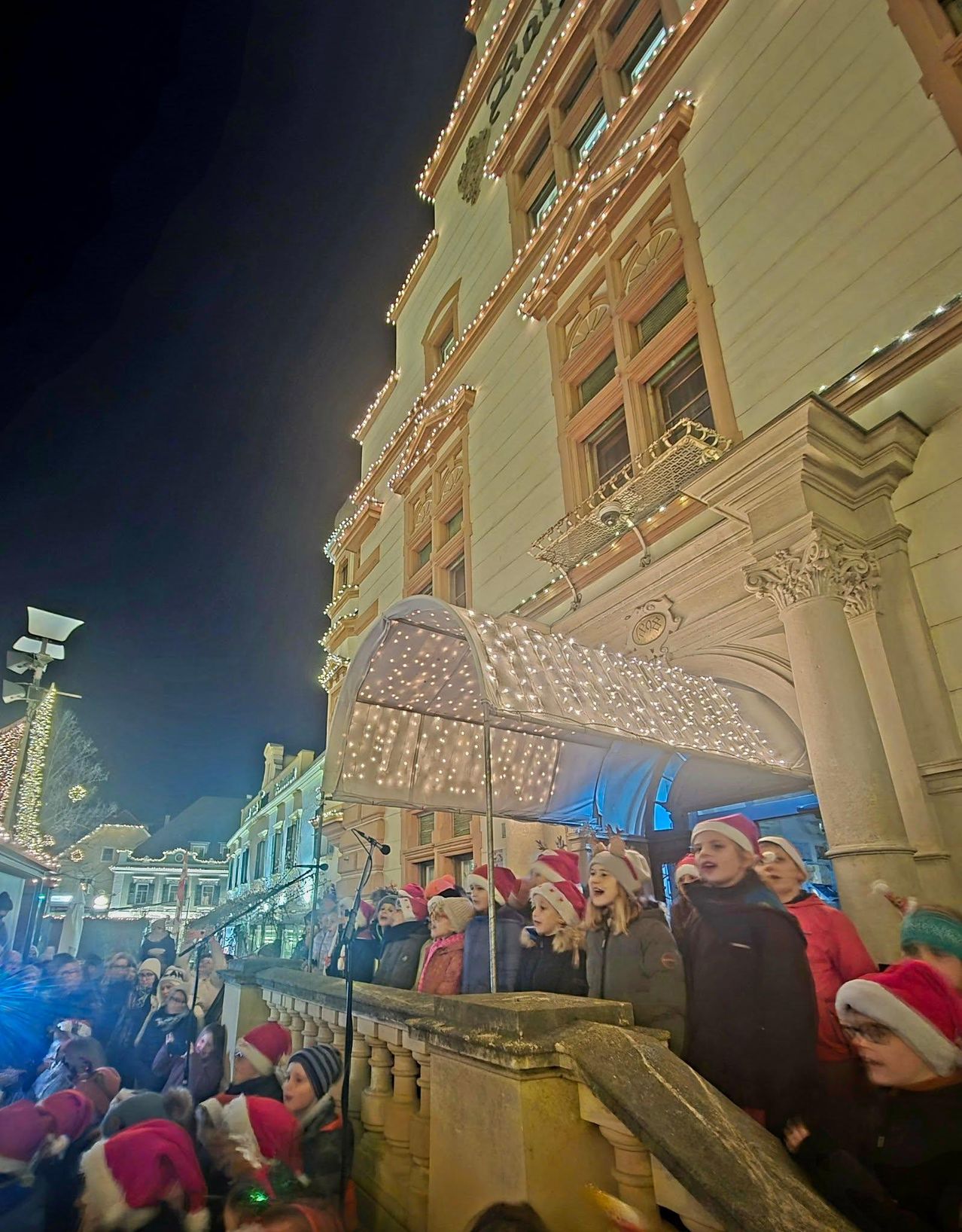 Ein Chor in Winterkleidung singt auf einer Bühne vor einem dekorierten Gebäude bei Nacht. Das Publikum, ebenfalls mit Mützen, beobachtet von unten.