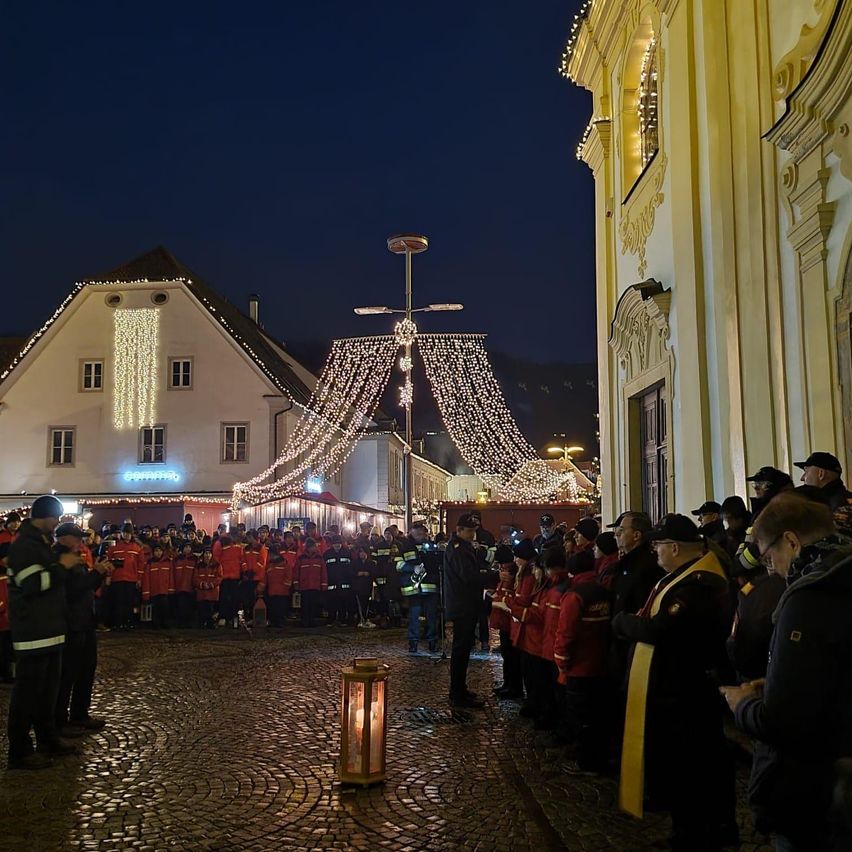 Bei Nacht steht eine Gruppe von Menschen in roten Jacken um eine Laterne auf einem Kopfsteinpflasterplatz, mit einem Gebäude und Weihnachtslichtern im Hintergrund.