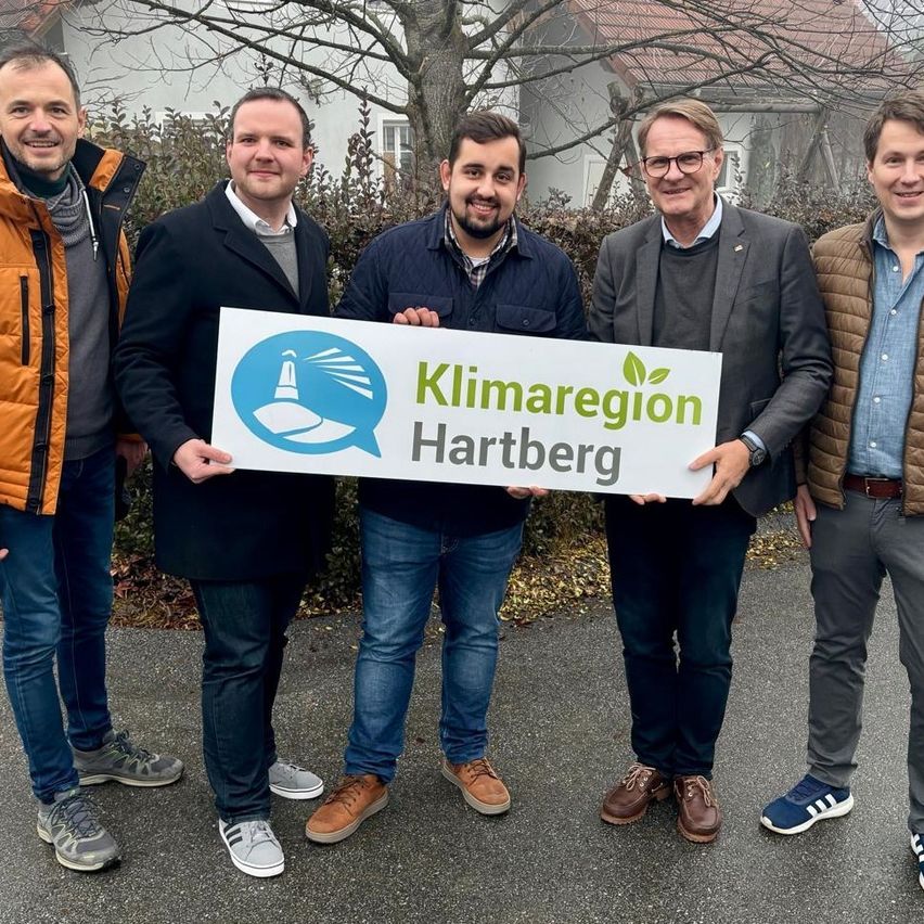Fünf Männer posieren mit einem Schild, das Klimaregion Hartberg steht vor einem Haus mit kahlen Bäumen.